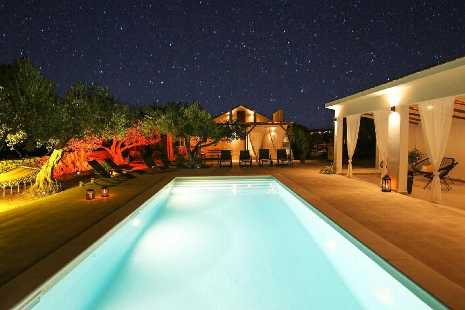 A swimming pool at night with a starry sky, a house, and outdoor seating.
