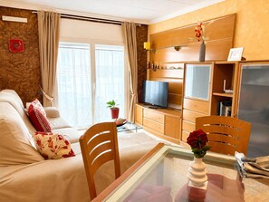 Una sala de estar con sofá, televisor, aparador de madera y una mesa de vidrio con un florero.