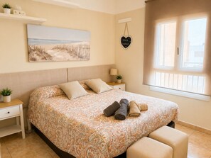 Un dormitorio con una colcha estampada, una mesita de noche con una lámpara, una planta junto a la cama y un cuadro con motivos playeros en la pared.