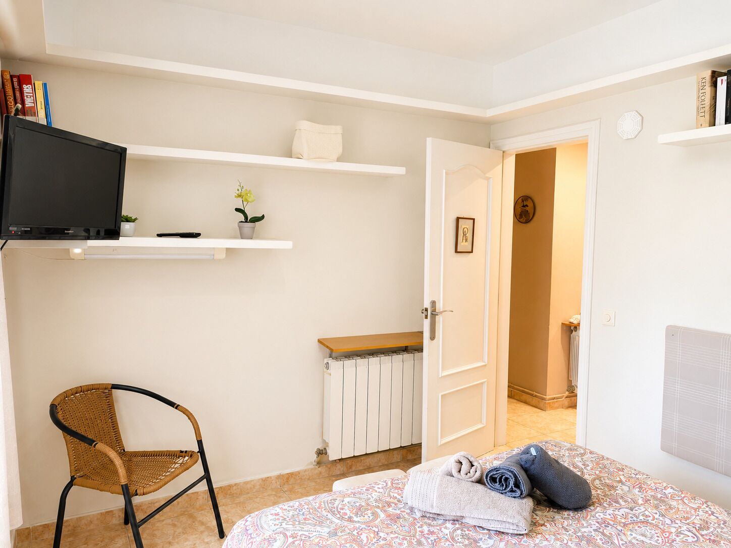 A bedroom with a bed, a chair, a TV, a radiator, and a door leading to another room.