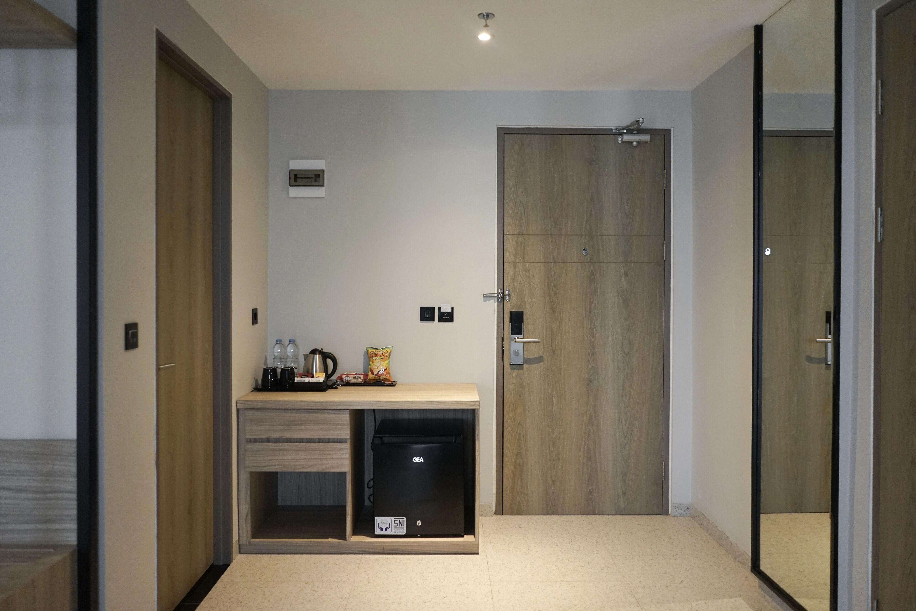 A modern hotel room with a wooden door, a minibar, a small trash bin, and a light fixture on the ceiling.
