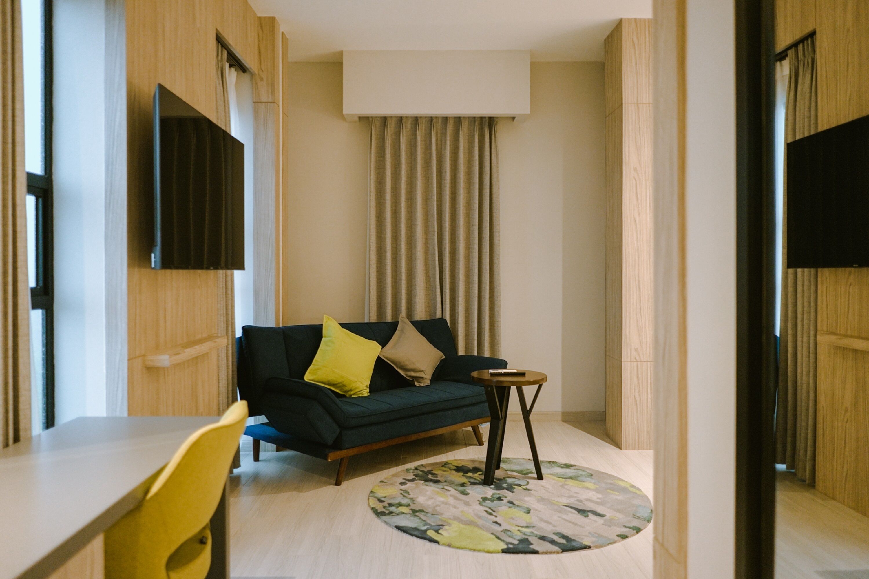 Una habitación de hotel moderna con un sofá verde oscuro, cojines amarillos y beige, una mesita redonda y una alfombra circular.