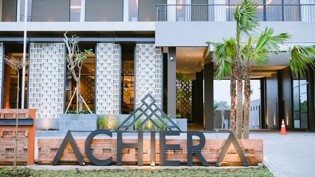 Un edificio moderno con una entrada destacada, un cartel decorativo y un área exterior bien cuidada con palmeras.