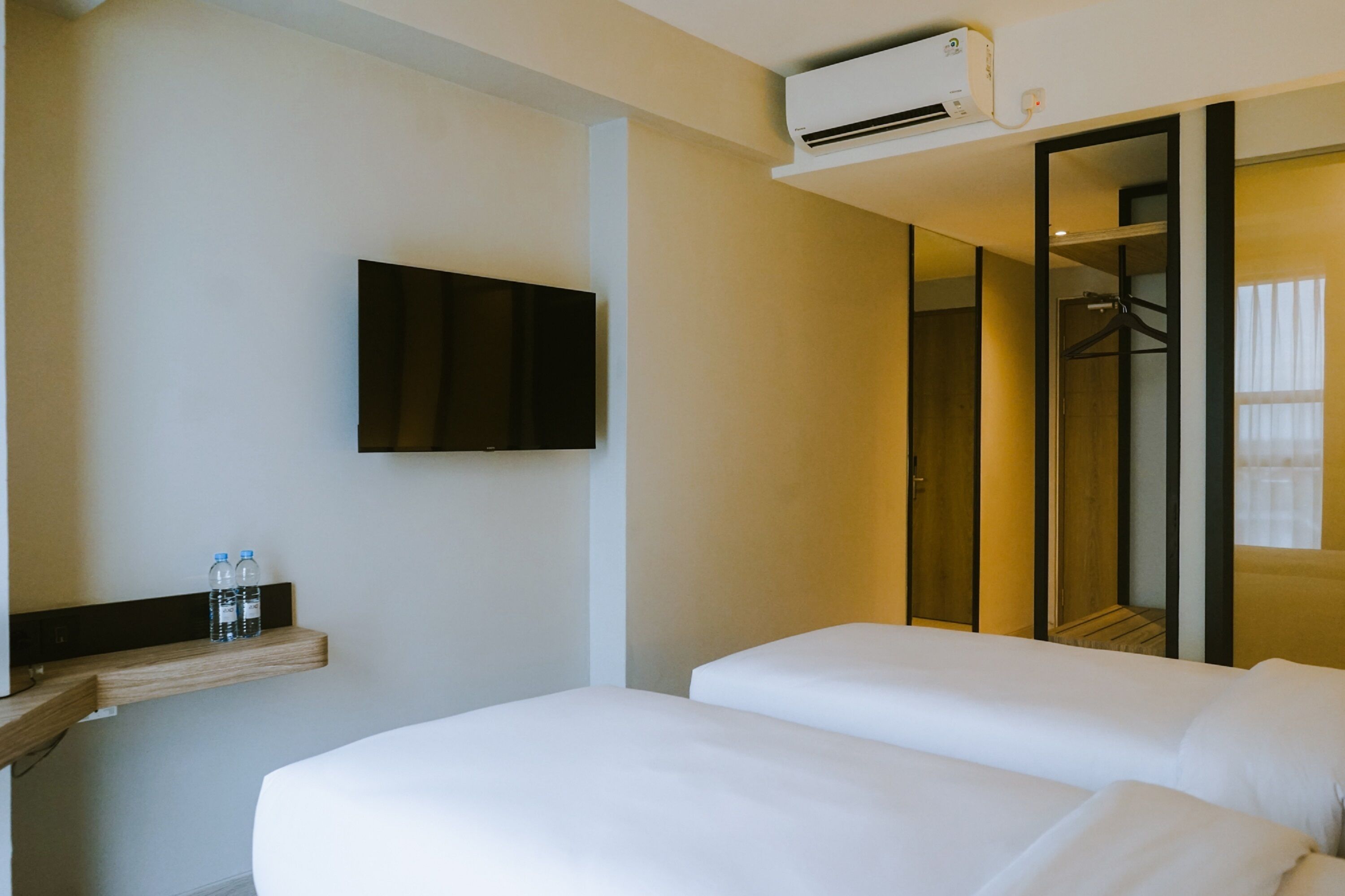 Habitación de hotel con dos camas, televisor montado, aire acondicionado y un espejo que refleja un armario y una ventana.