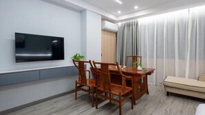 A modern hotel room with a wooden dining table, chairs, a flat-screen TV mounted on the wall, a sofa, and a large window with curtains.