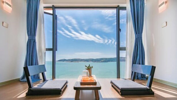 A room with a large window, two chairs, a small table, and a view of the sea. - Piaoyang Guohai Guesthouse (Zhoushan)