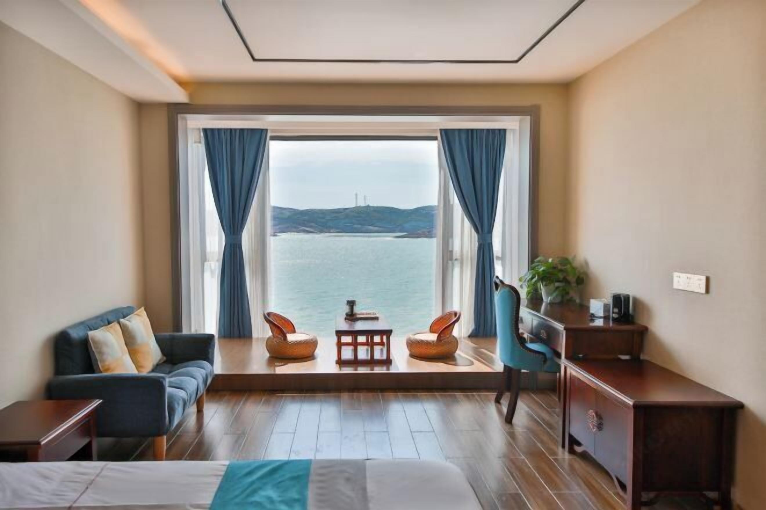 A room with a bed, a sofa, two chairs, a small table, and a desk. A large window offers a view of the sea and distant hills.