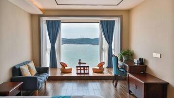 A room with a bed, a sofa, two chairs, a small table, and a desk. A large window offers a view of the sea and distant hills.