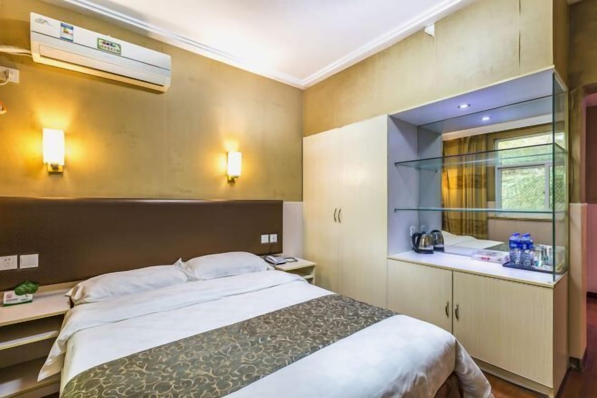 A hotel room with a bed, bedside table, lamp, wardrobe, and a window with a view.