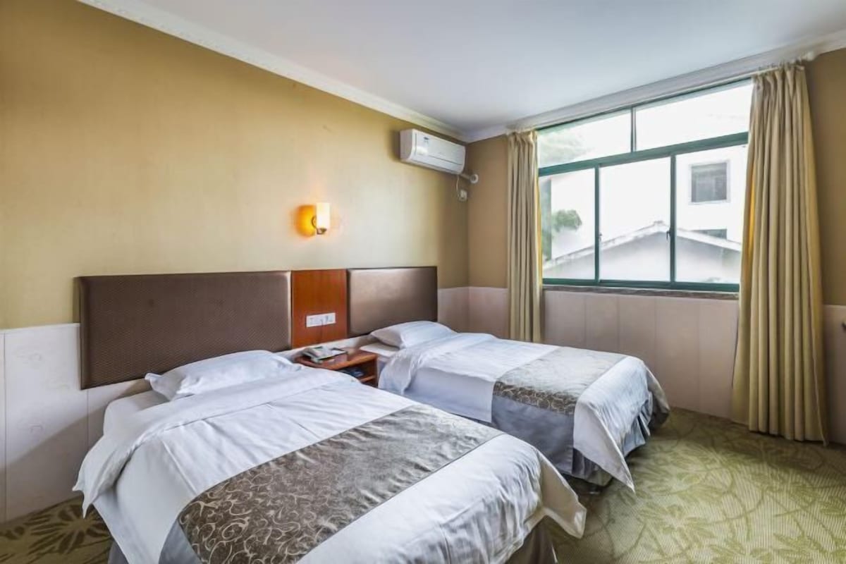 A hotel room with two beds, a wall-mounted air conditioner, a nightstand, and a window with curtains.