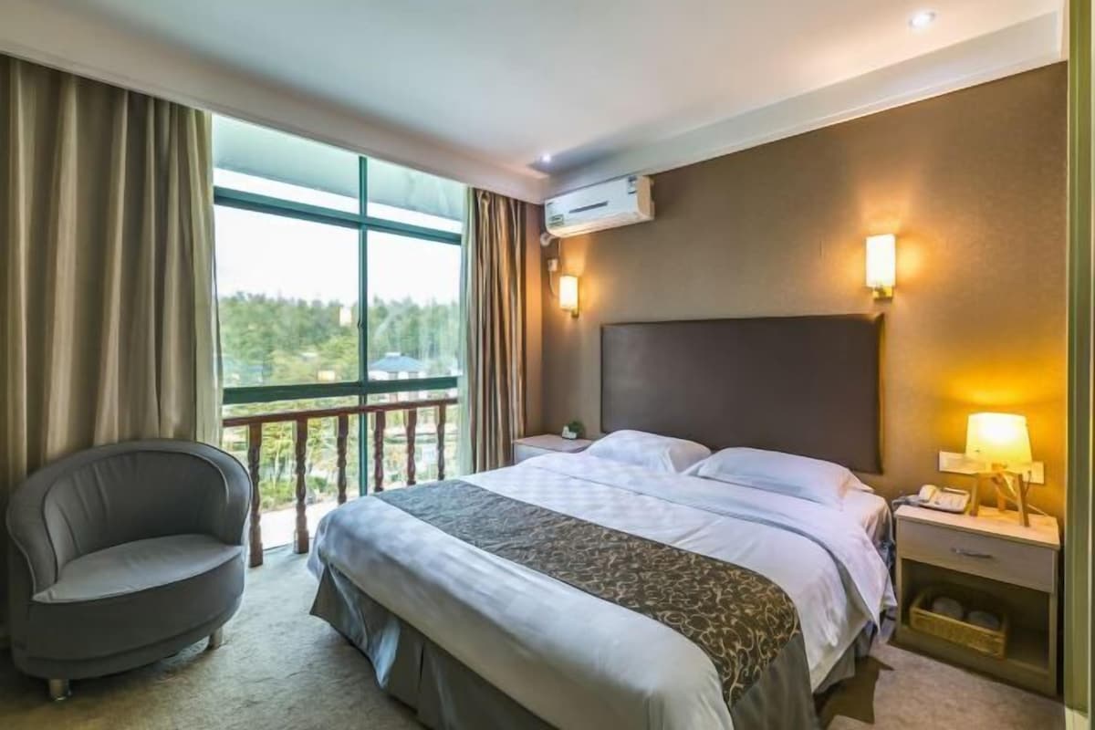 A hotel room with a large bed, a chair, a nightstand with a lamp, and a view of greenery outside.