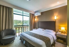 Habitación de hotel con una cama grande, una silla, una mesita de noche con lámpara y vistas a la vegetación.