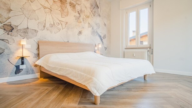 A bedroom with a wooden bed, a stone wall with a leaf pattern, and a window with a view of buildings. - The Roman Villa View - Italian Homing (Desenzano del Garda)