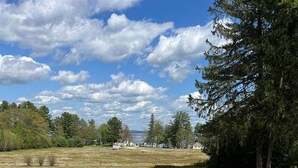 A grassy outdoor area with two lounge chairs facing a fire pit and a view of a lake and trees.