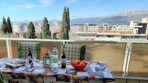 Un balcón con una mesa dispuesta para una comida, que incluye una botella de vino, copas y un bol de naranjas. El fondo muestra edificios y montañas.