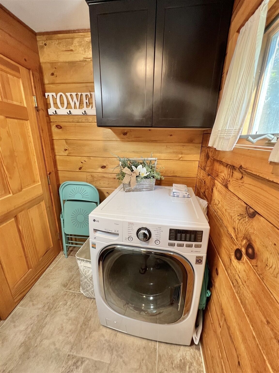 A compact laundry room with a washing machine and dryer, wooden walls, a small chair, and a cabinet above.