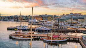 Un puerto deportivo con varias embarcaciones amarradas, un horizonte urbano al fondo y un cielo al atardecer.
