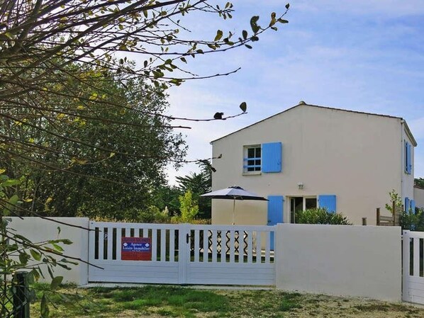 A two-story building with a white exterior and blue shutters, surrounded by a white fence with a sign indicating it's for sale.