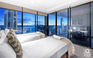 Enjoy a panoramic view of the city skyline and ocean from the large balcony, featuring two beds with white linens and a glass partition.