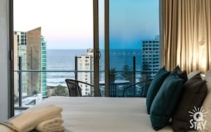 Enjoy ocean and city views from the large window in this inviting room, featuring a comfortable bed and a small table with a chair.