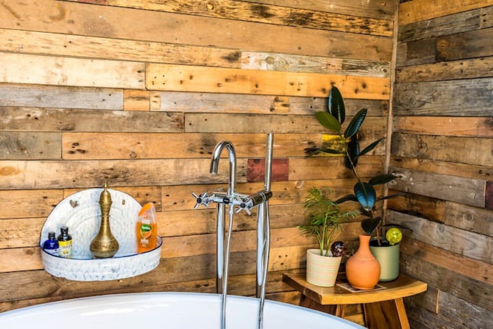 Une salle de bain avec un mur en bois, un lavabo et un miroir. On y trouve des articles de toilette sur une étagère et une plante en pot sur une petite table.