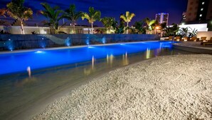 A swimming pool with a pebble beach area, palm trees, and a cityscape in the background. - 2 Bedroom Ocean View Apartment - Q Stay (Surfers Paradise)