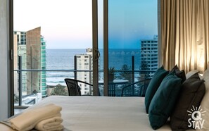 Enjoy ocean and city views from the large window in this inviting room, featuring a comfortable bed and a small table with a chair.