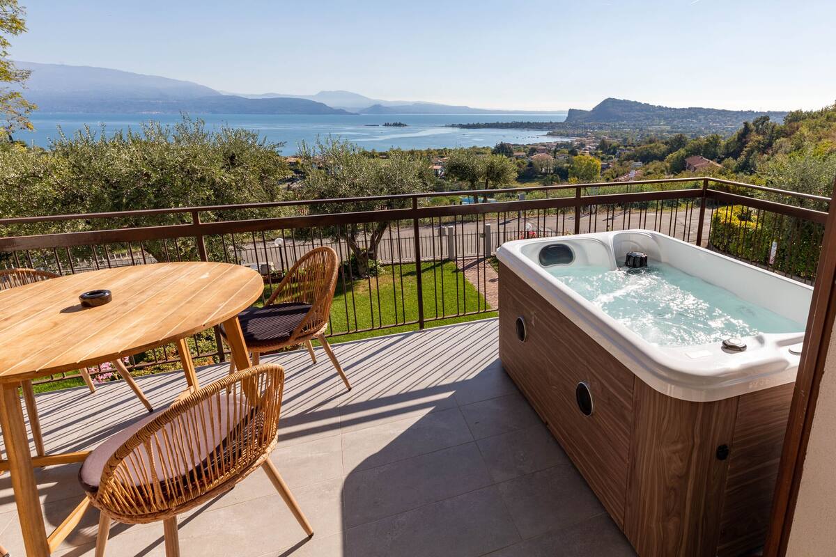 A balcony with a wooden table and chairs, a hot tub, and a scenic view of a lake and mountains.