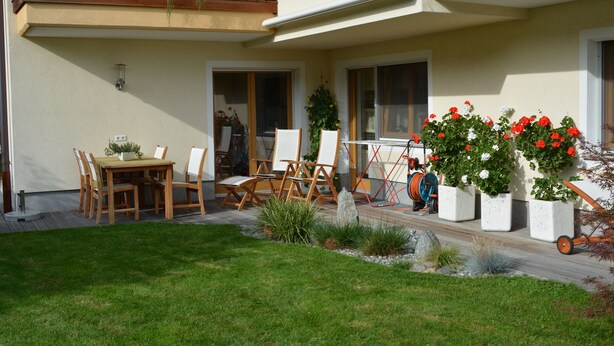 A residential building with a patio area featuring a dining table and chairs, a small garden with potted plants, and a wooden deck. - Apartment with beautiful terrace to relax (Kapfing)