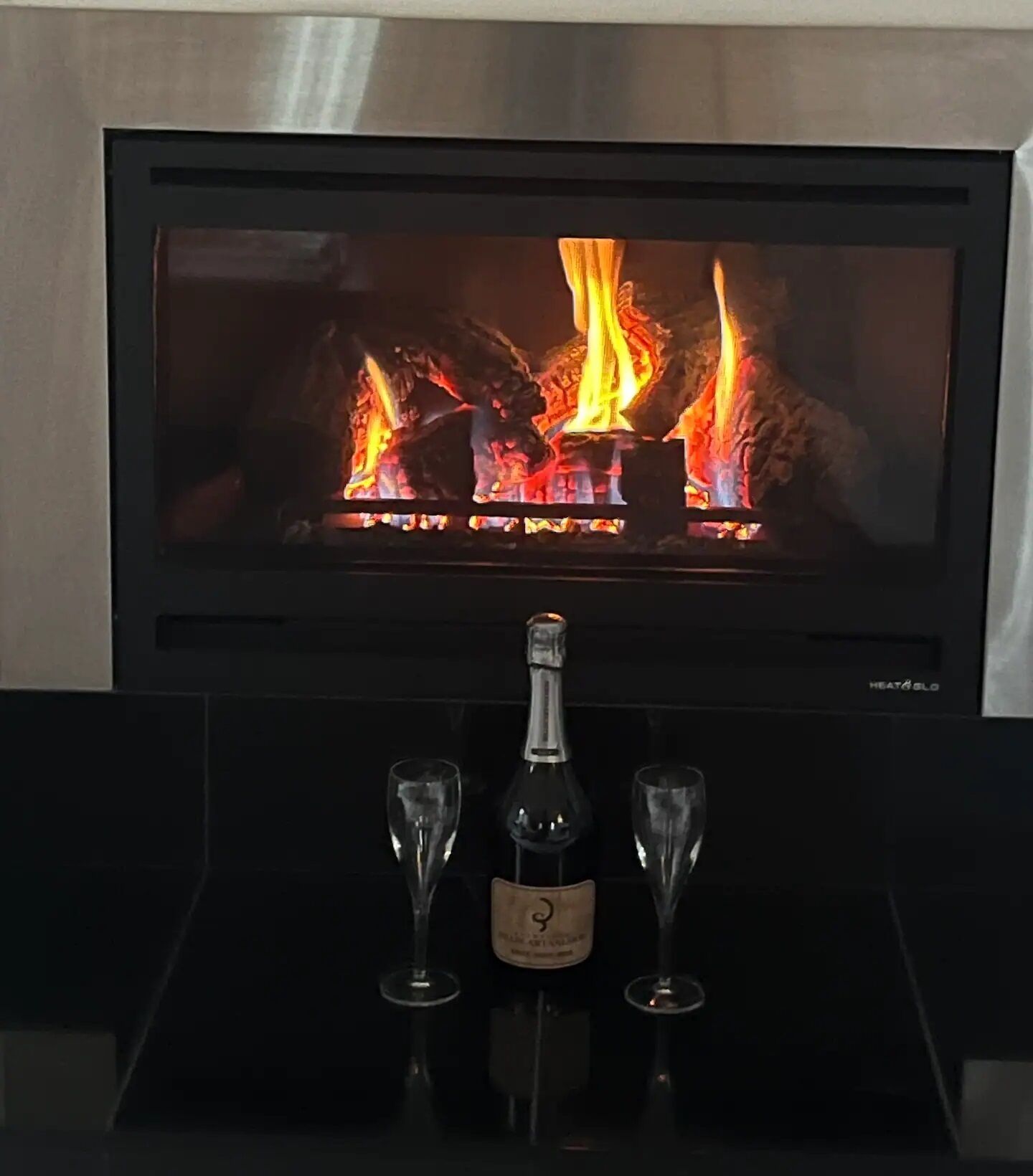 Ein Kamin mit Fernseher, der ein Feuer zeigt, eine Weinflasche und zwei Weingläser auf einem schwarzen Tablett.