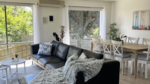A modern living room with a black leather sofa, a dining table with white chairs, and large windows offering a view of greenery outside. - Cosy Family Home 500 metres from Red Rock Beach (Cowes)