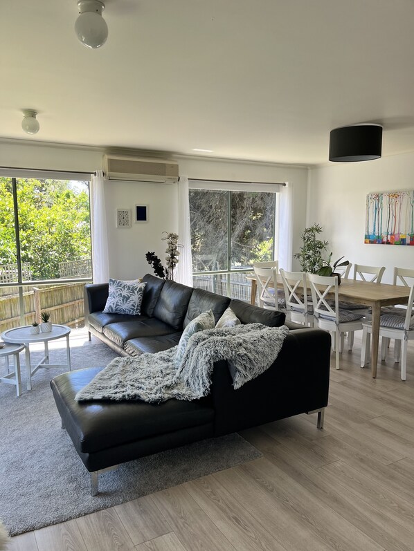 A modern living room with a black leather sofa, a dining table with white chairs, and large windows offering a view of greenery outside. - Cosy Family Home 500 metres from Red Rock Beach (Cowes)