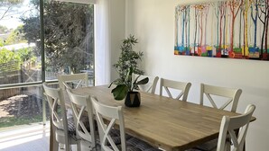 A dining area with a wooden table and white chairs, a potted plant, and a colorful painting on the wall. - Cosy Family Home 500 metres from Red Rock Beach (Cowes)