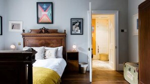 Une chambre à coucher avec un lit en bois, des tables de chevet avec des lampes, une porte donnant sur un couloir et un tableau encadré au mur.