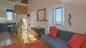 A spacious living area with a grey sofa, red cushions, a wooden dining table, and a kitchen in the background. - Alfama, charming attic Terrace with river view - Lisboa vida boa 1  (Lisboa)