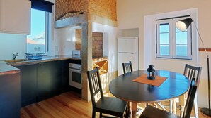 A modern kitchen with a brick accent wall, a dining area with a round table and chairs, and a view of the sea through the windows. - Alfama, charming attic Terrace with river view - Lisboa vida boa 1  (Lisboa)