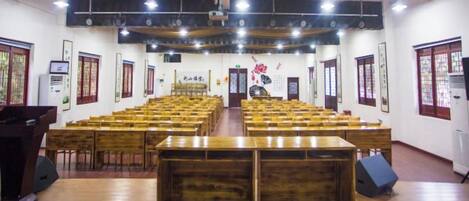 Un salón amplio con filas de mesas y sillas de madera, un escenario en la parte delantera y decoraciones chinas tradicionales.