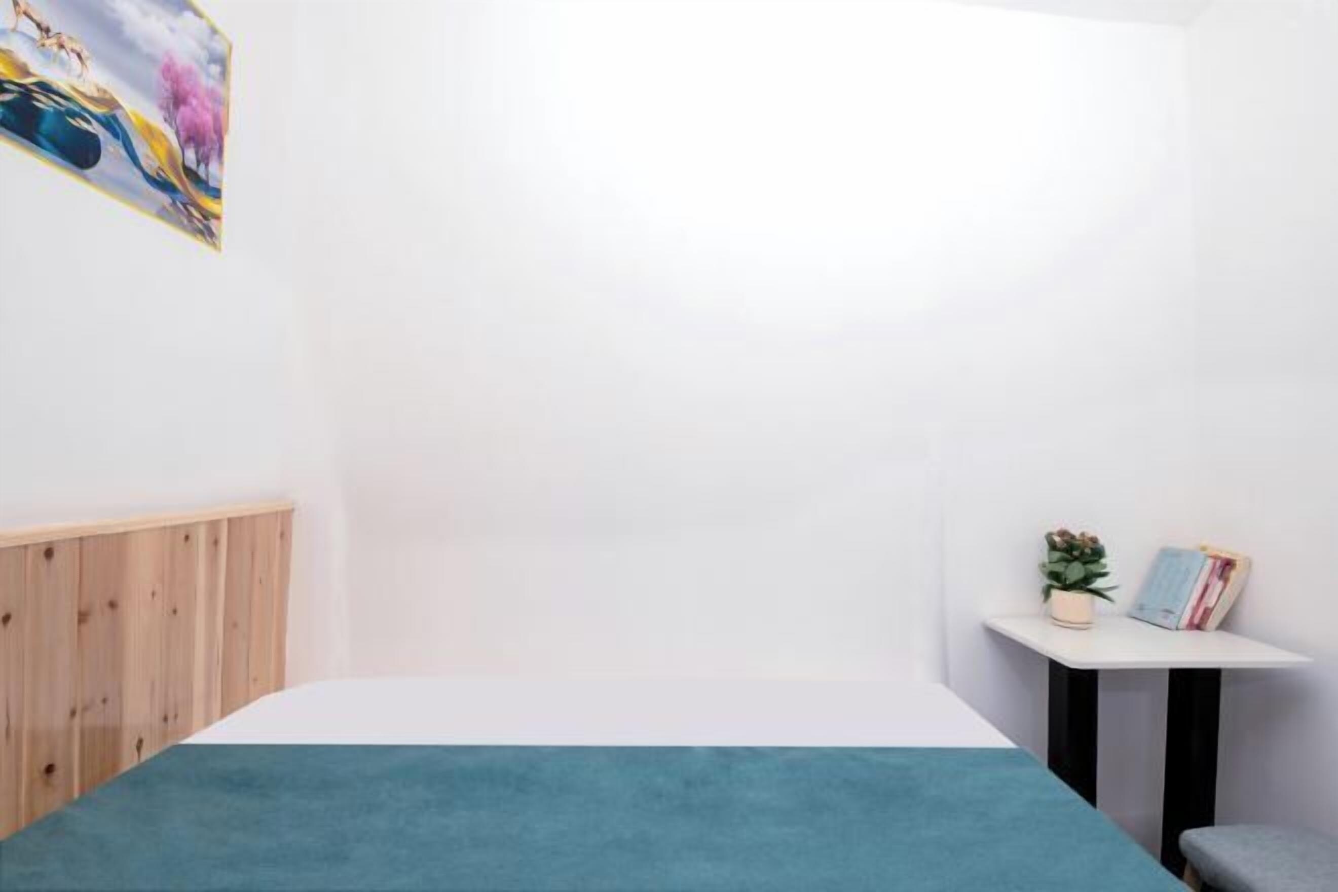 Une chambre à coucher avec un lit, une tête de lit en bois, une petite table avec une plante et des livres, et un tableau accroché au mur.