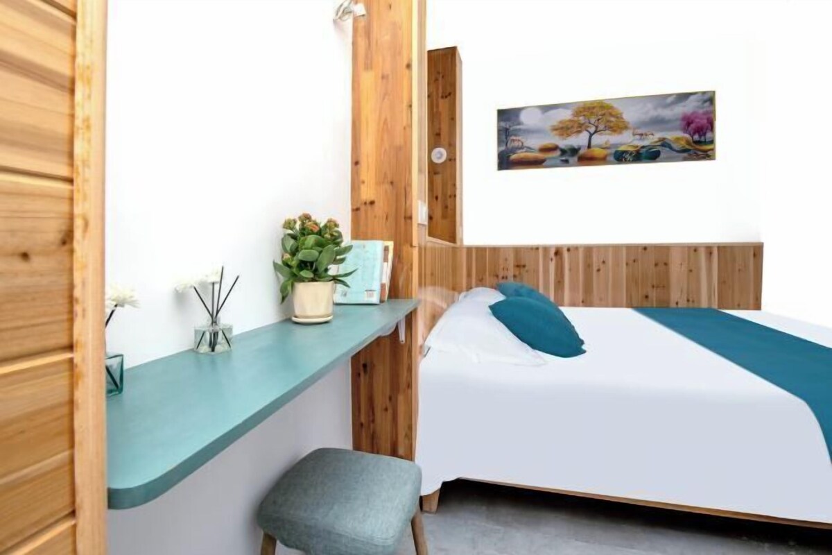 A bedroom with a bed, a desk, a chair, a potted plant, and a painting on the wall.