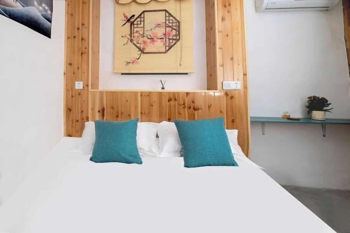 A bedroom with a wooden headboard, a bed with white bedding and teal pillows, a wall-mounted artwork, and a small shelf with a plant.