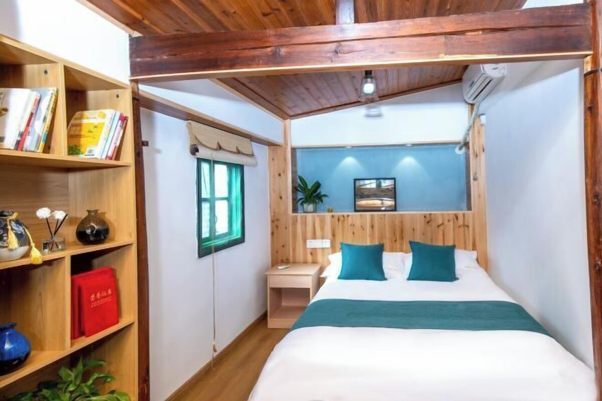 A bedroom with a wooden headboard, a bed with teal pillows, a bedside table, a nightstand, a bookshelf with books and decorative items, and a window with a curtain.