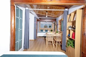 A cozy room with a wooden ceiling, a bed with a blue headboard, a wooden dining table with chairs, and a bookshelf.