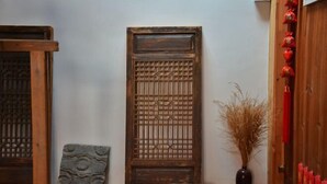 Une porte en bois traditionnelle ornée de sculptures complexes, une plaque de pierre avec un motif circulaire et un vase contenant des plantes séchées.