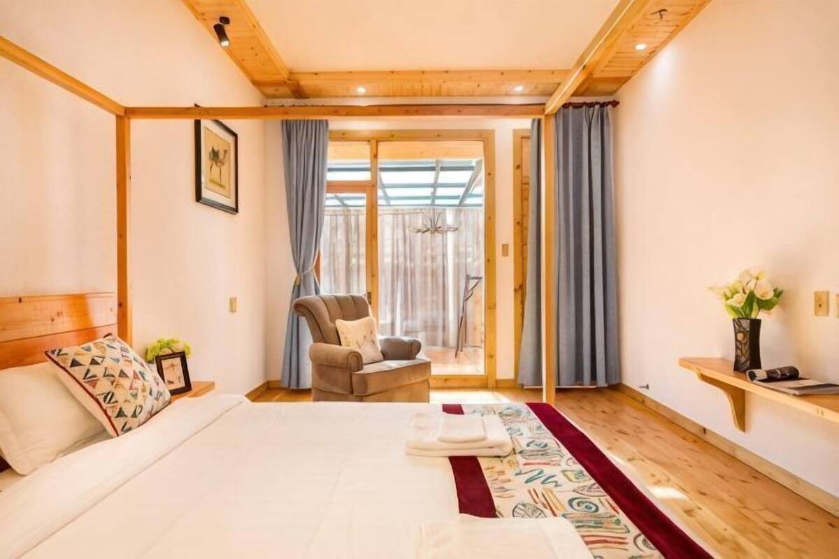 Une chambre à coucher avec un lit, une chaise, une petite table sur laquelle se trouve un vase, et une vue à travers une porte vitrée.