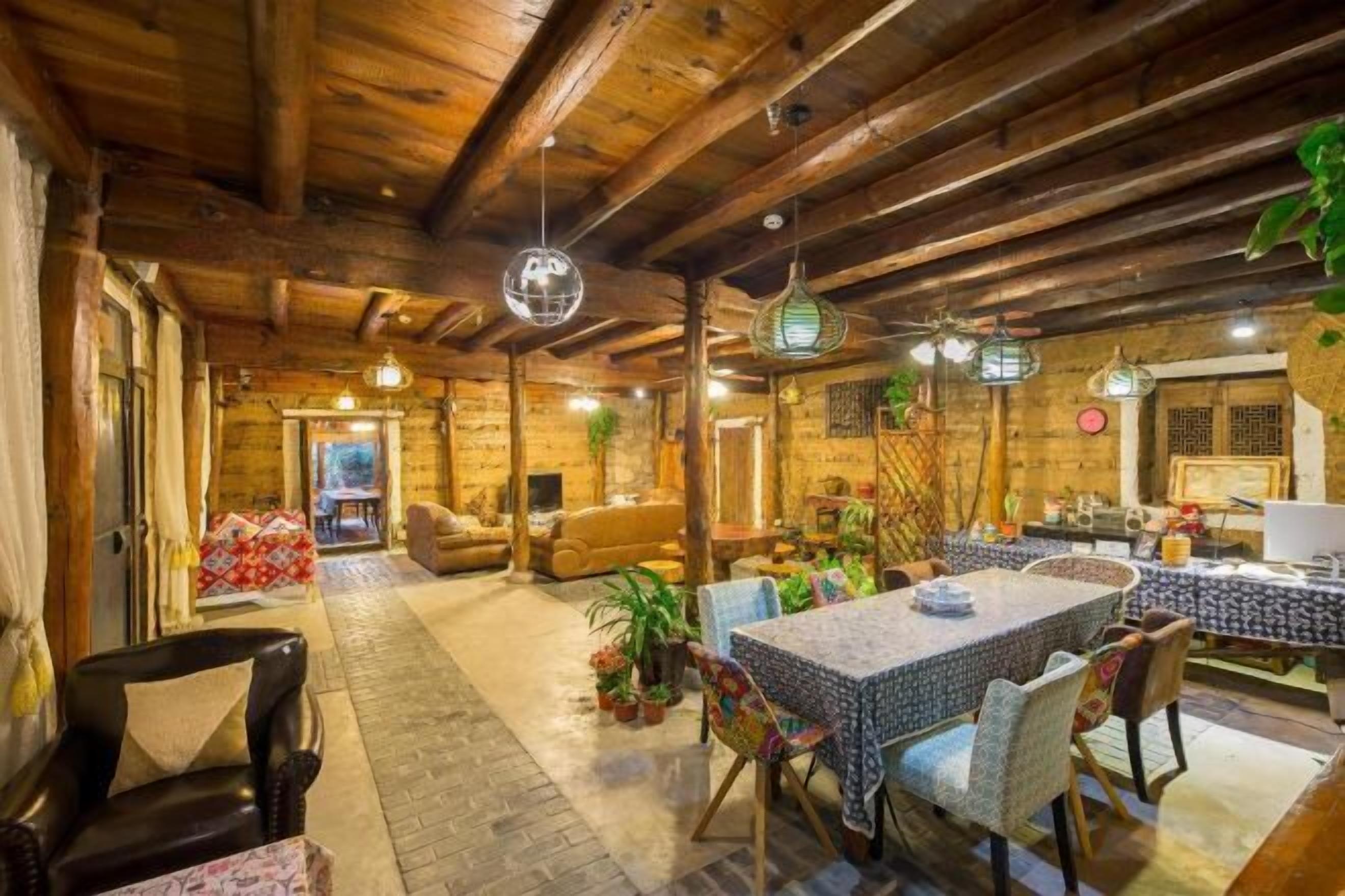 A rustic interior with exposed wooden beams, a dining area with a patterned tablecloth, and various plants.