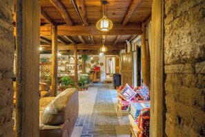 A narrow corridor with a stone wall on one side and a wooden ceiling with hanging lights. There is a leather sofa and a small table with items on the left, and a display of colorful textiles on the right.
