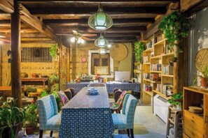 A dining area with a long table, chairs, hanging lamps, and bookshelves filled with books and plants.