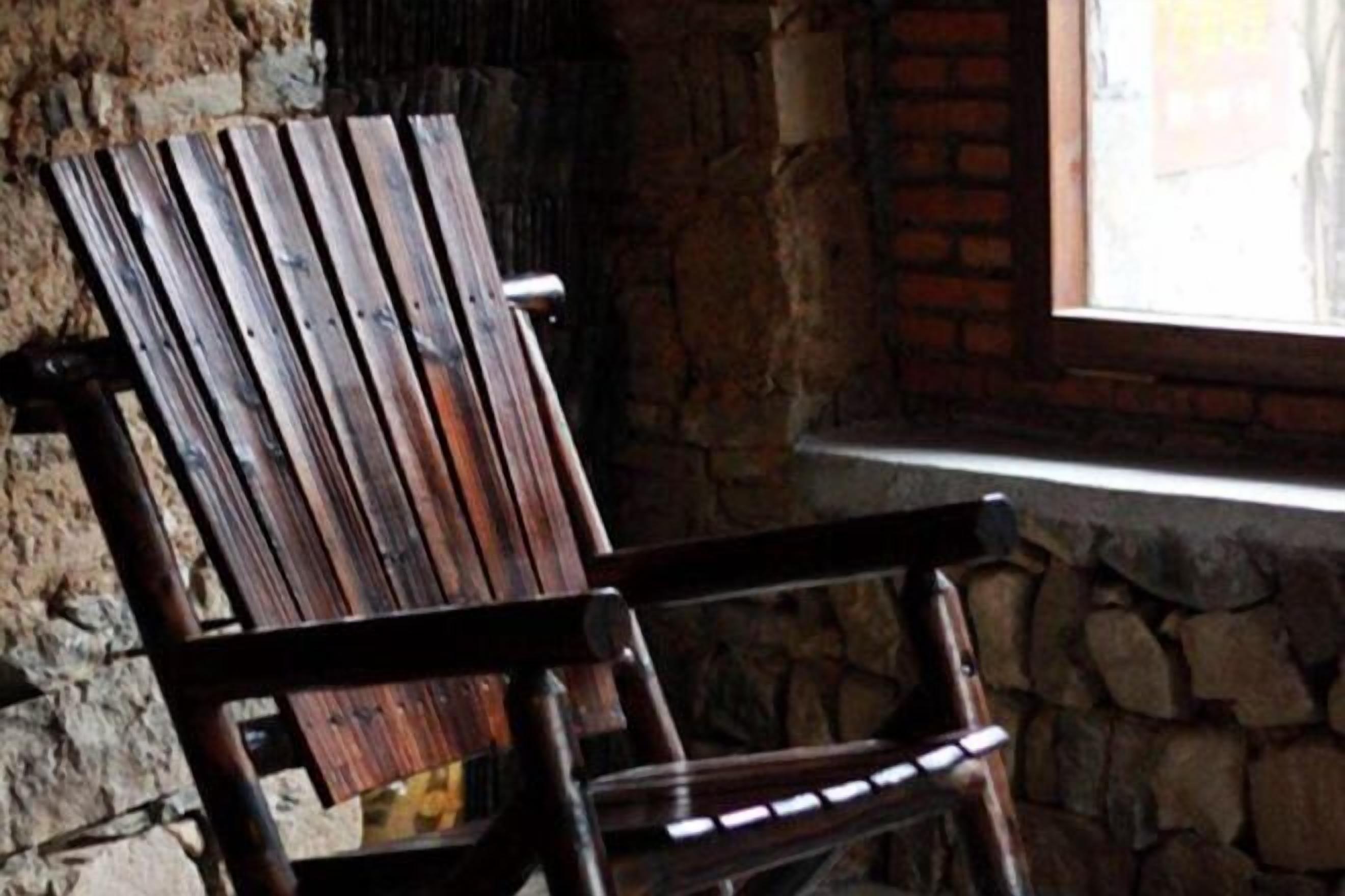 A rustic wooden chair with a slatted backrest, positioned against a stone wall with a window above it.