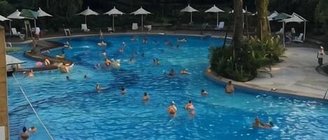 A swimming pool with multiple people, surrounded by umbrellas and lounge chairs.
