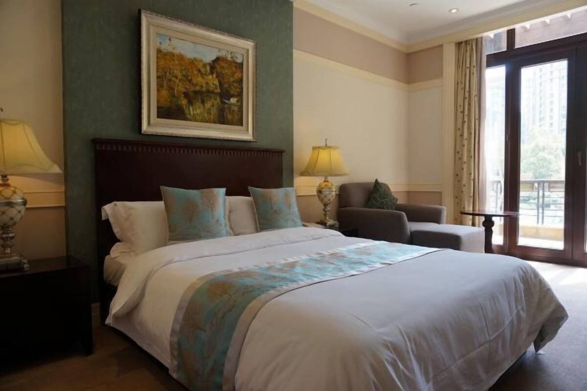 A hotel room with a large bed, two bedside tables with lamps, a chair, a painting on the wall, and a balcony with curtains.